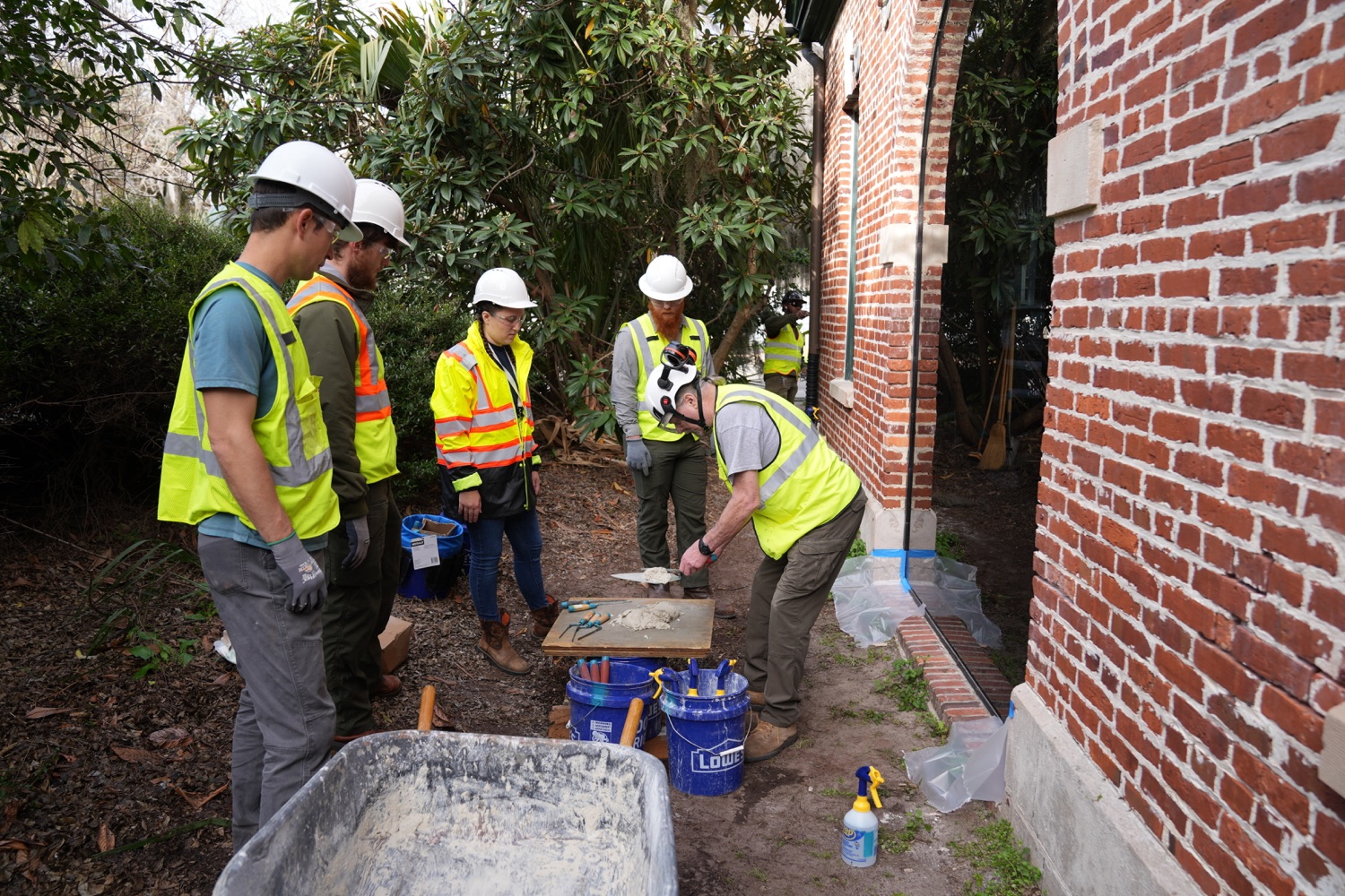 National Park Service craftspeople demonstrating mortar preparation and brick repointing beside a historic brick wall.
